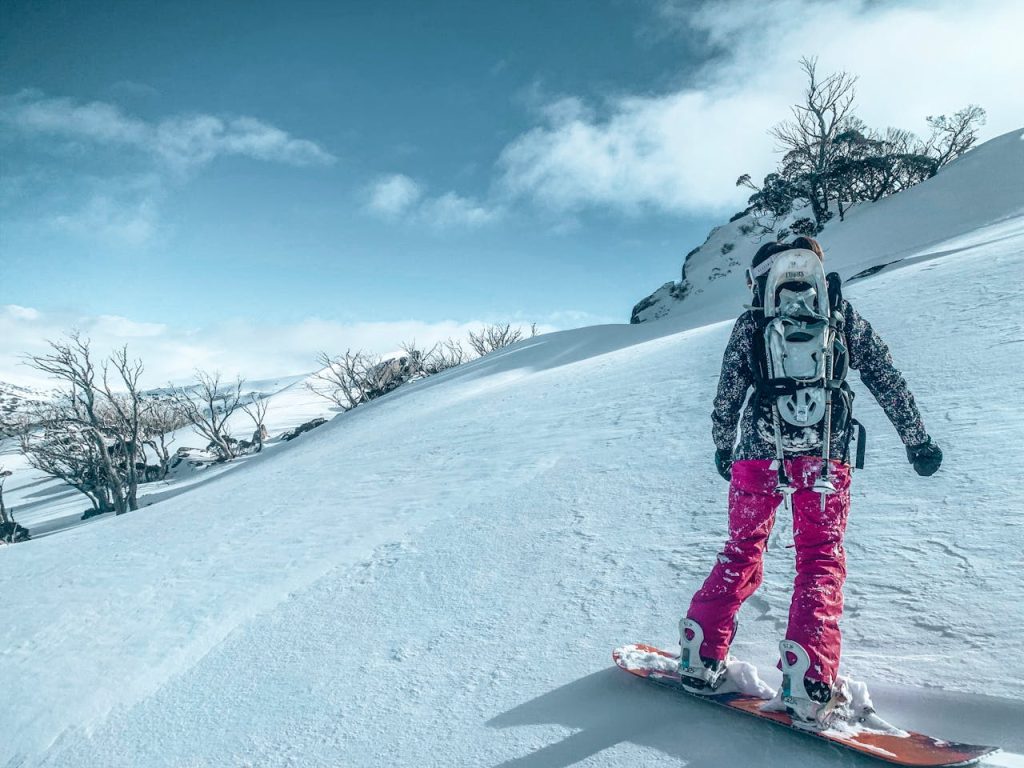 Person in Black Jacket and Pink Pants Standing on Snow Covered Ground Snowboarding